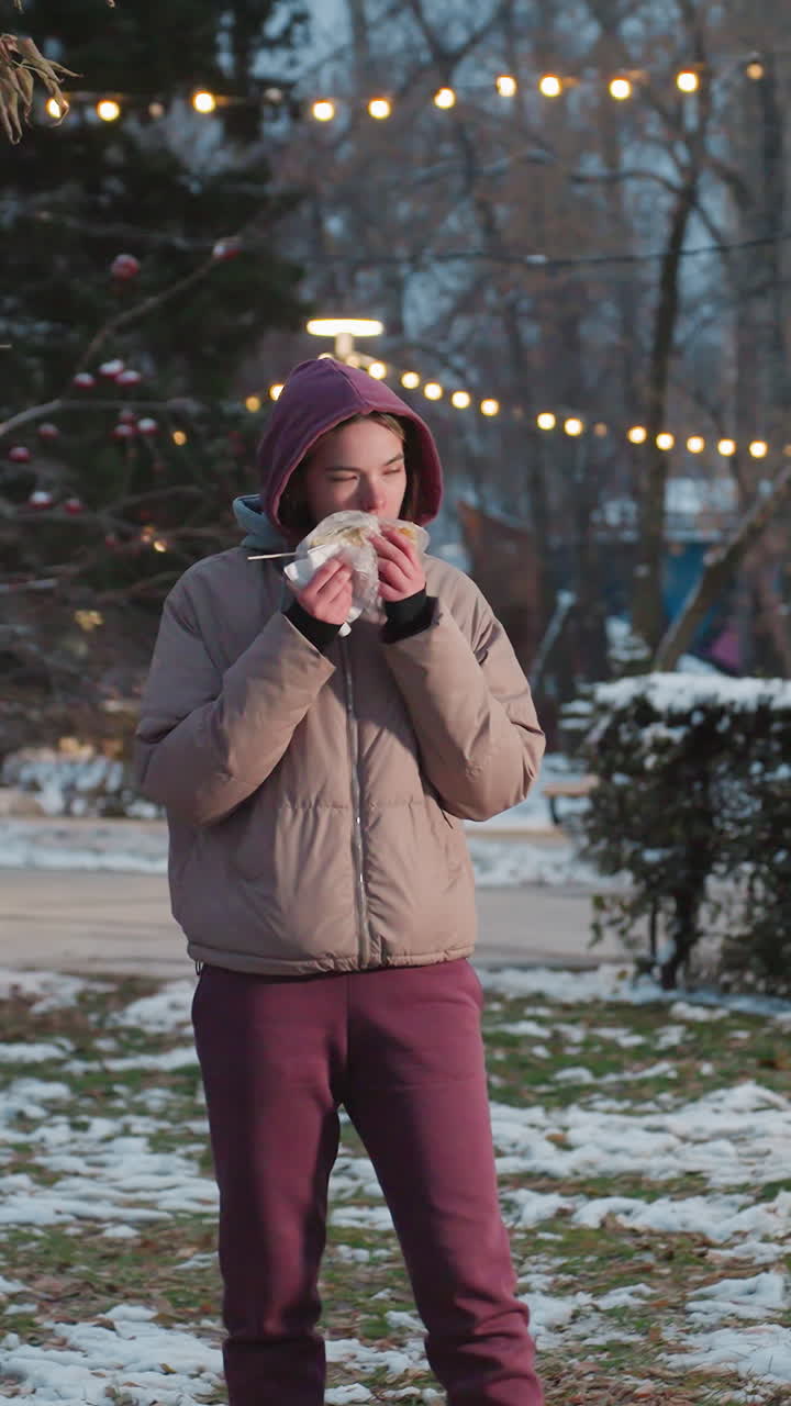 joven de pie en el parque sosteniendo una servilleta mientras come maíz, escena de invierno al aire libre con nieve en el suelo, bokeh de luz cálida en el fondo