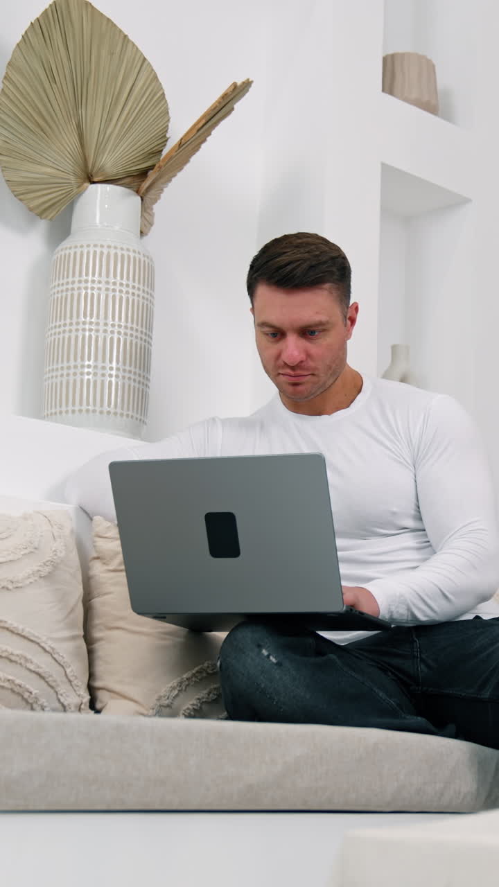 Dark-haired Caucasian male sits on sofa focused on laptop. Working remote from home. Low angle view. Vertical video