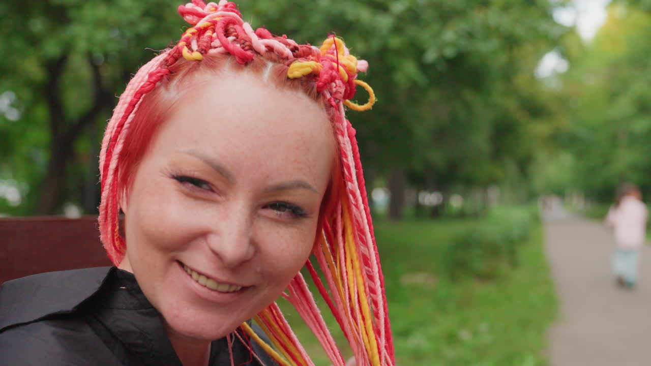 햇빛 아래에서 분홍색과 노란색 땋은 머리를 한 쾌활한 여성이 행복하게 웃고 있는 모습, 생동감 넘치는 땋은 머리를 한 표현력 풍부한 여성이 야외에 앉아 미소를 지으며 입을 가리고 있어요.