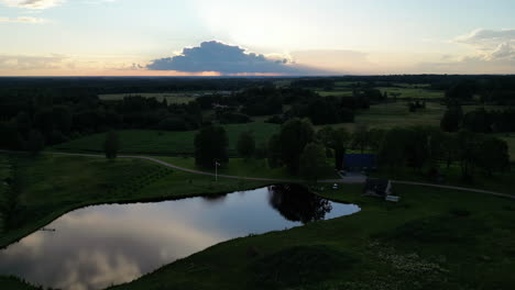 Time-lapse-of-a-sunset,-sun-hiding-behind-the-cloud,-with-green-fields-and-trees-and-a-small-lake-nearby-a-single-family-house,-copy-space