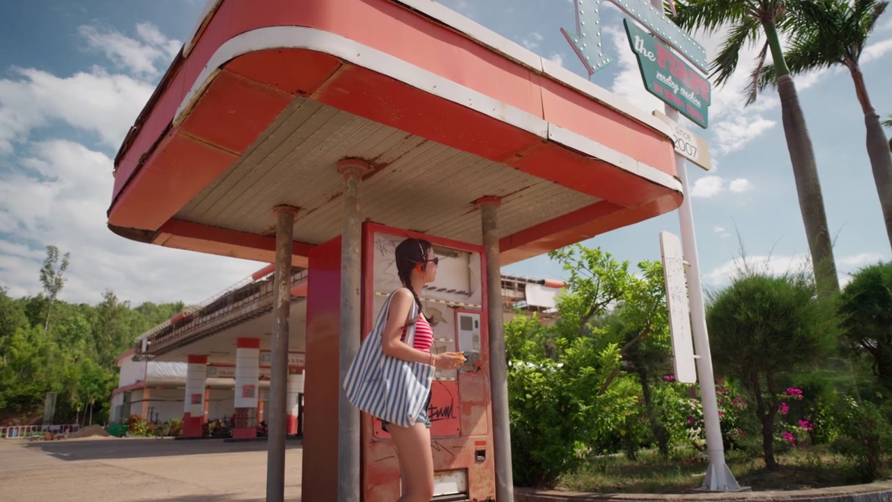 Giovane donna balla mentre ondeggia ascoltando un riproduttore di cassette portatile accanto a un distributore automatico rovinato dalle intemperie con segni di graffiti all'aperto, un lento allontanamento rivela una stazione di servizio.