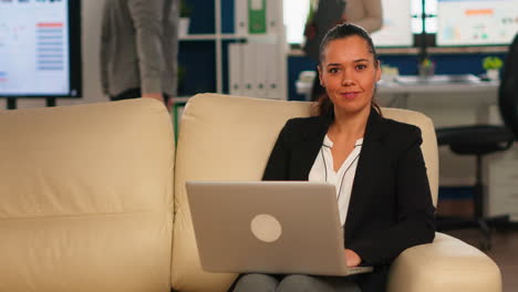 Hispanische-Geschäftsfrau-Lächelt-In-Die-Kamera,-Sitzt-Auf-Der-Couch-Und-Tippt-Am-Computer
