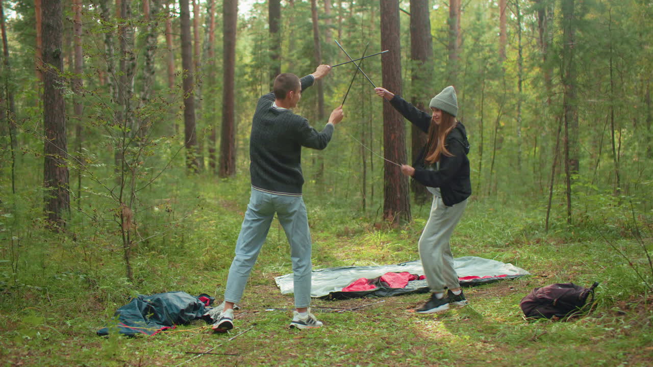 Cheerful couple in forest playfully spar with tent poles, mimicking karate moves while preparing camp, surrounded by lush greenery and scattered camping gear during lighthearted outdoor moment