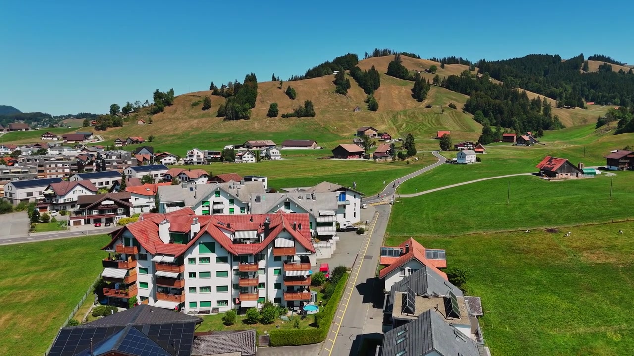Aerial view near Sihlsee in Switzerland, showing an Alpine village with apartment buildings, green meadows, winding roads and rolling farmland beneath forested hills.