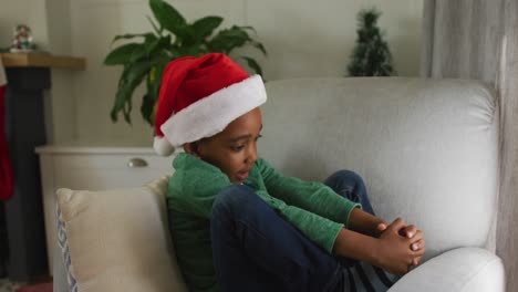 Sad-african-american-boy-wearing-santa-hat-sitting-on-sofa-at-christmas-time