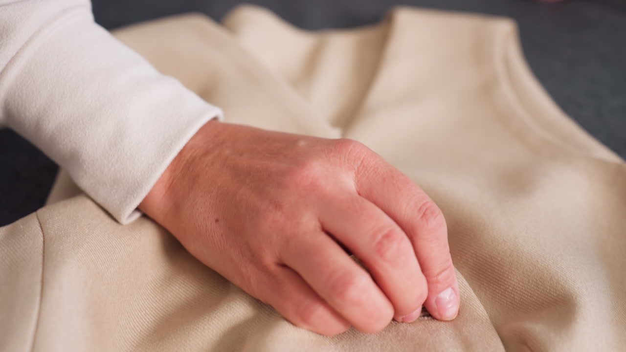 Dress maker carefully removes buttons from beige winter jacket placed on dark table, using fingers to gently detach fasteners while preserving fabric texture and garment details