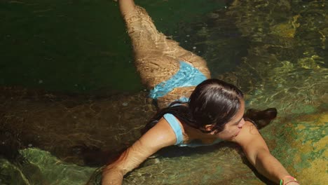 young latina relax in a blue bikini in the clear pool at a waterfall on a sunny day