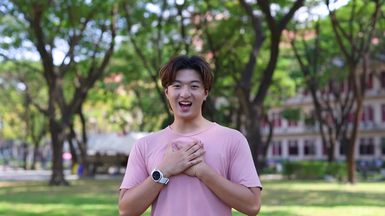 Seorang pria muda Asia berkaus merah muda mengungkapkan rasa terima kasih yang tulus di taman yang cerah