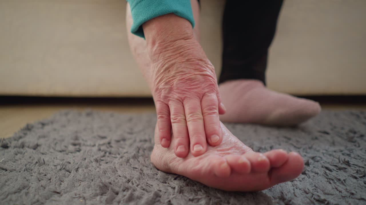 Old woman in green applying ointment to her ankle while seated on bed in cozy indoor environment. She appears focused, taking care of herself, possibly for pain or discomfort relief