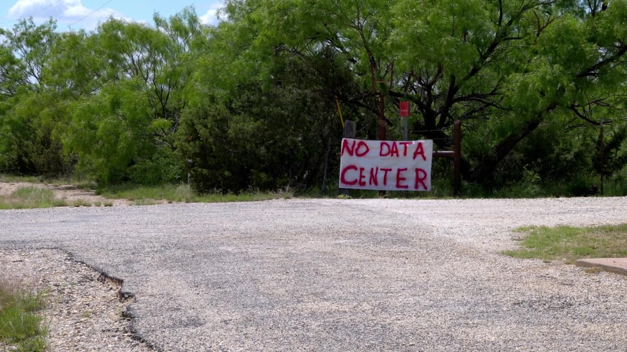 Uma placa branca caseira, pintada em vermelho com 'No Data Center', destaca-se contra um cenário de árvores verdes exuberantes. Ela significa resistência da comunidade local e preocupações ambientais em relação a propostas.