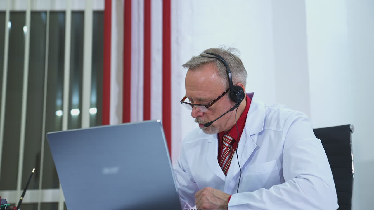 Telemedicine. Old physician listens to a patient from laptop. Male doctor wear headset make online video call consultation to a patient in clinic.