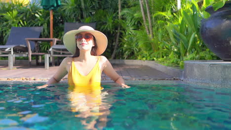 Una-Mujer-Joven-Y-Bonita-Con-Traje-De-Baño-Amarillo,-Gafas-De-Sol-Y-Un-Sombrero-Para-El-Sol-Se-Inclina-A-Lo-Largo-Del-Borde-Trasero-De-Una-Piscina-Del-Complejo