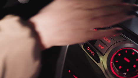 vista en primera persona de la mano de una mujer presionando el botón del mapa en un sistema de navegación de coche por la noche. el tablero está iluminado con luces rojas, destacando los controles. disparado con una cámara de mano