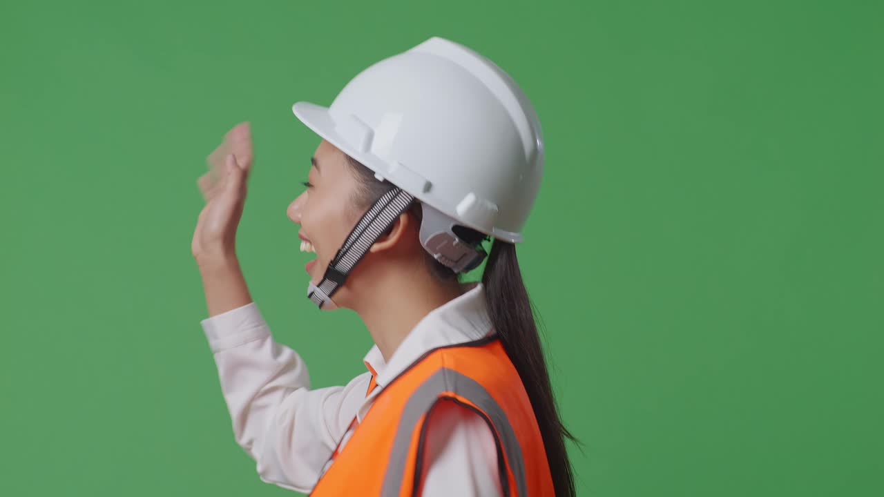으로 가게 볼 때, 안전 헬을 입은 아시아 여성 엔지니어가 녹색 스크린 배경 스튜디오에서 는 동안 손을 흔들고 있습니다.