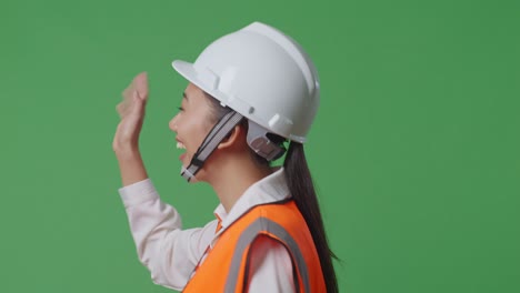 vista lateral de cerca ingeniera asiática con casco de seguridad agitando la mano mientras camina en el estudio de fondo de la pantalla verde