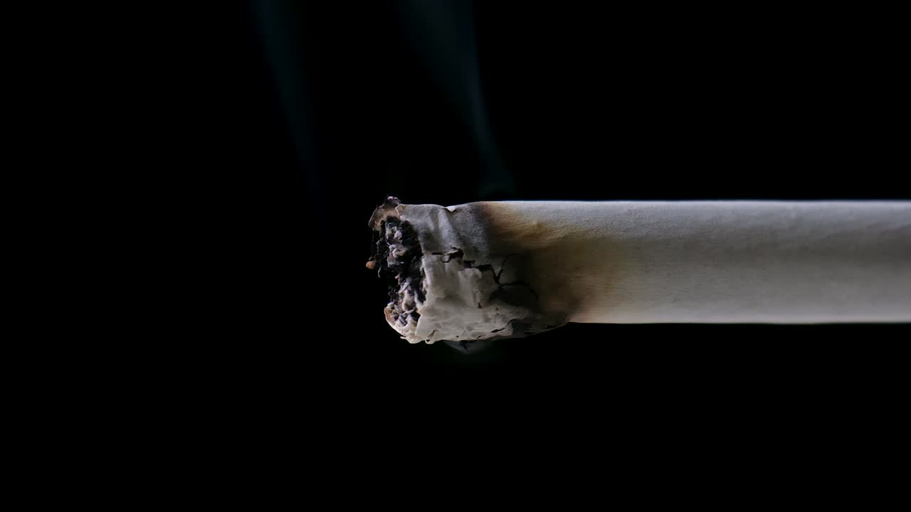 zoom en la toma de la punta de un cigarrillo consumiéndose lentamente, emitiendo un rastro de humo, frente a un fondo negro