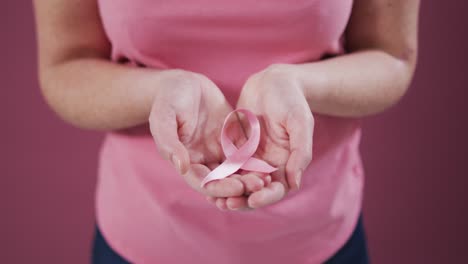 Sección-Media-De-Una-Mujer-Con-Las-Manos-Ahuecadas-Sosteniendo-Una-Cinta-Rosa-De-Concientización-Sobre-El-Cáncer-De-Mama.