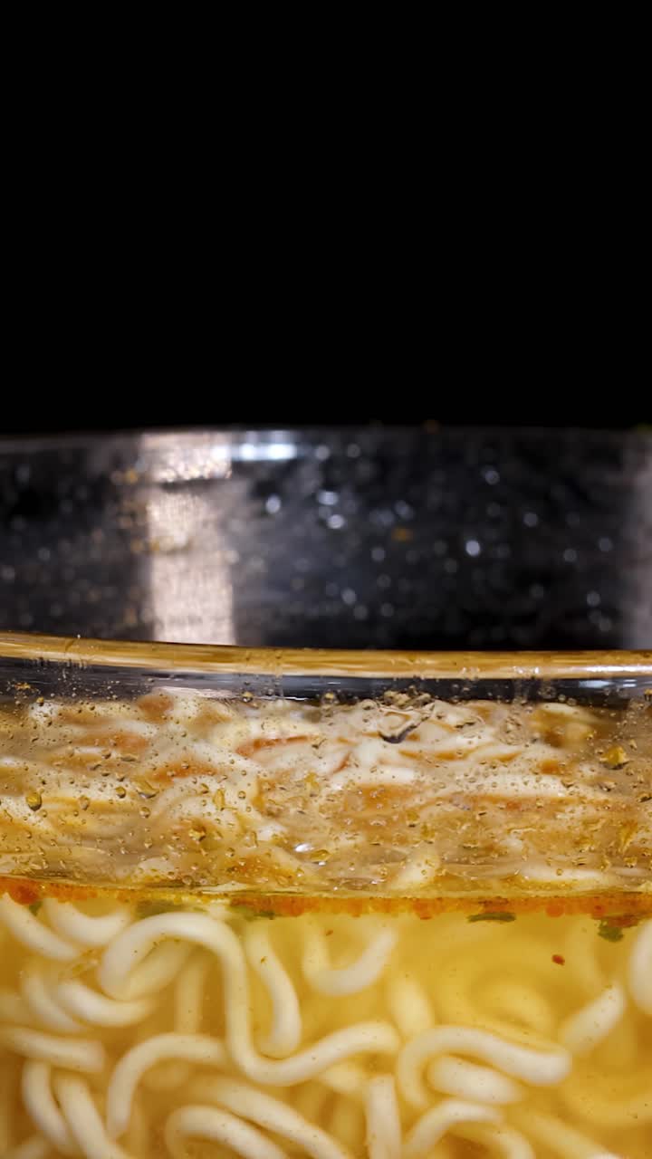 Video macro di una ciotola di vetro con noodles istantanei bollenti, erbe aromatiche e brodo in rotazione sotto una luce intensa