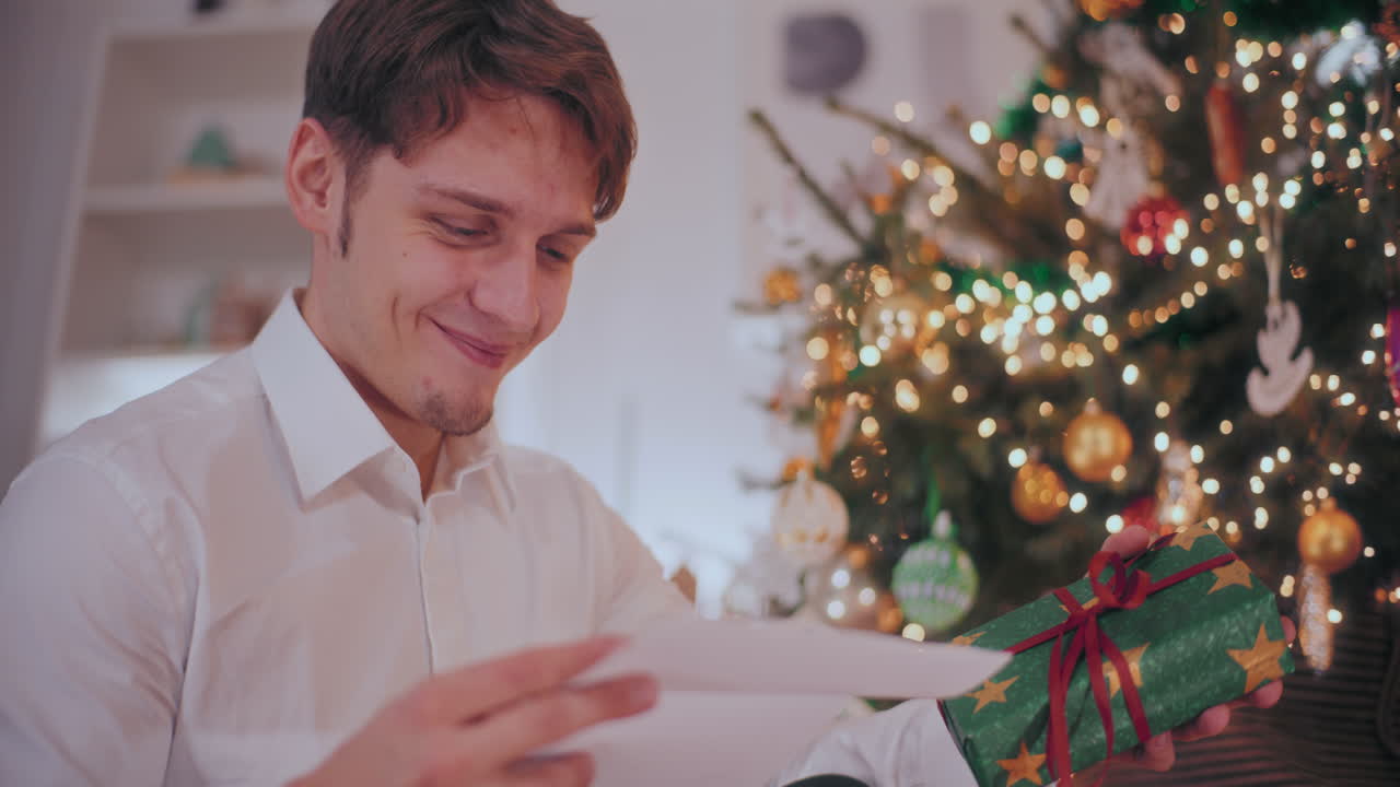 hombre sonriente sosteniendo un regalo de navidad mientras lee una carta en casa