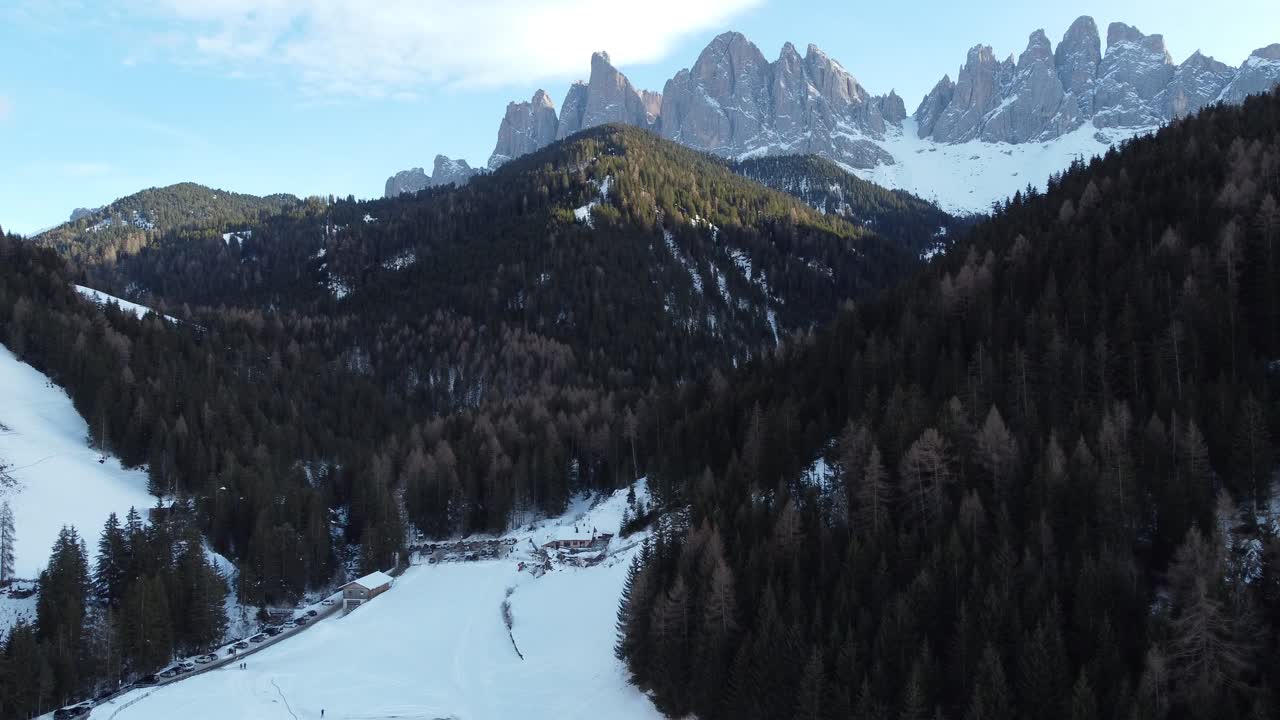 Snow-covered valley surrounded by dense pine forests and dramatic jagged peaks under blue sky in Trentino-Alto Adige captured from above during serene winter day