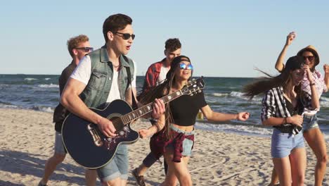 group of young hipster friends walking and dancing together playing guitar and singing songs on a beach at the water's edge. slowmotion shot