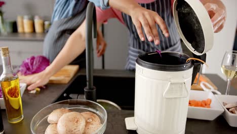 Midsection-of-diverse-couple-in-aprons-preparing-vegetables-composting-waste-in-kitchen,-slow-motion