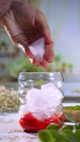 hand dropping ice cubes into a curvy glass with strawberry mash at the bottom, preparing a vibrant iced base for a matcha latte or coffee strawberry drink in a bright, natural setting.