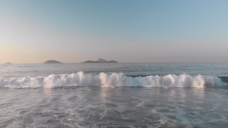 Early-morning-golden-hour-hovering-low-over-water-backing-up-with-a-small-breaking-ocean-wave-coming-in-at-sunrise-until-reaching-the-beach-with-islands-in-the-background