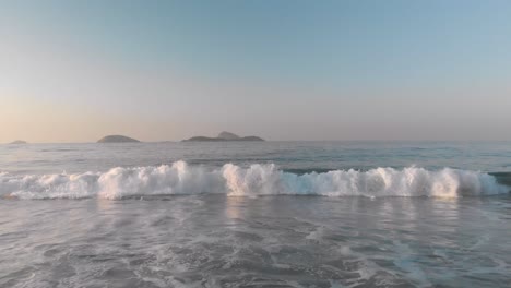 early morning golden hour hovering low over water backing up with a small breaking ocean wave coming in at sunrise until reaching the beach with islands in the background