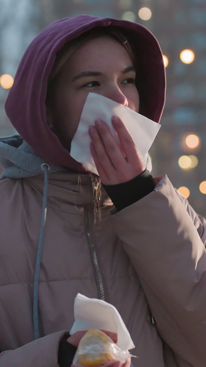 mujer sosteniendo un bocadillo con una servilleta en la mano limpiando la boca en un entorno al aire libre con luces bokeh en el fondo, temporada de invierno frío, disfrutando de la comida callejera, limpiándose la boca después de comer un delicioso bocadillo