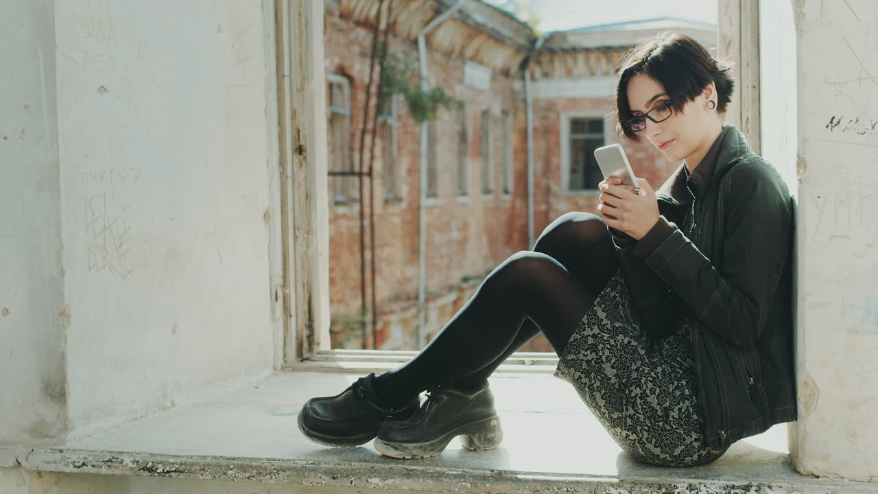 joven hipster sentada en el alféizar de una ventana en un edificio antiguo usando un teléfono celular