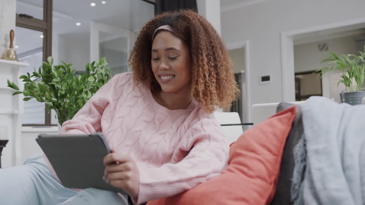 Happy biracial woman relaxing on couch using tablet at home, in slow motion