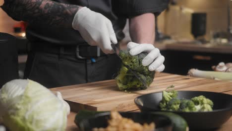 Fresh-green-broccoli-being-cut-on-a-wooden-board-by-young-professional-male-chef-in-an-elegant-black-shirt-with-tattoos