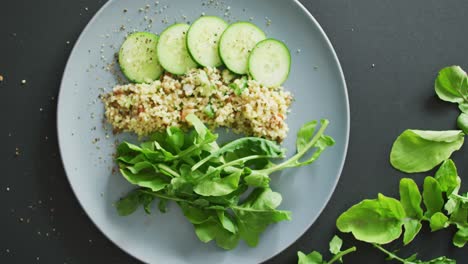 Video-of-fresh-sliced-cucumber,-leaves-and-grains-salad-on-grey-plate-over-dark-grey-background