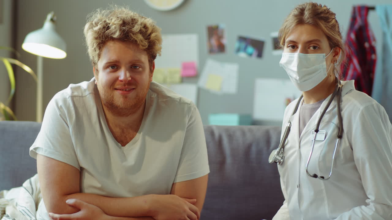Portrait of Patient and Doctor in Mask Sitting on Sofa at Home