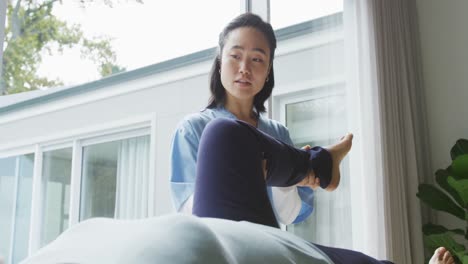 Asian-female-physiotherapist-holding-leg-of-female-patient-lying-on-examination-bed-at-surgery