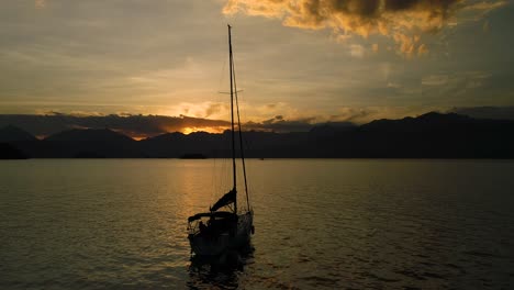 Hermoso-Dron-Aéreo-Orbita-Alrededor-De-Un-Velero-En-Un-Océano-Tranquilo-Con-Una-Increíble-Puesta-De-Sol-Naranja-Con-Nubes-Y-Montañas