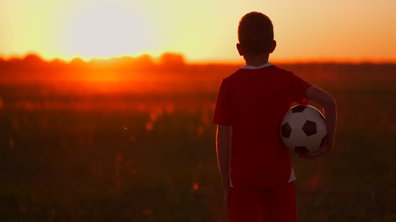 un niño con una pelota en un campo al atardecer. un niño sueña con convertirse en un jugador de fútbol. un niño va al campo con la pelota al anochecer.