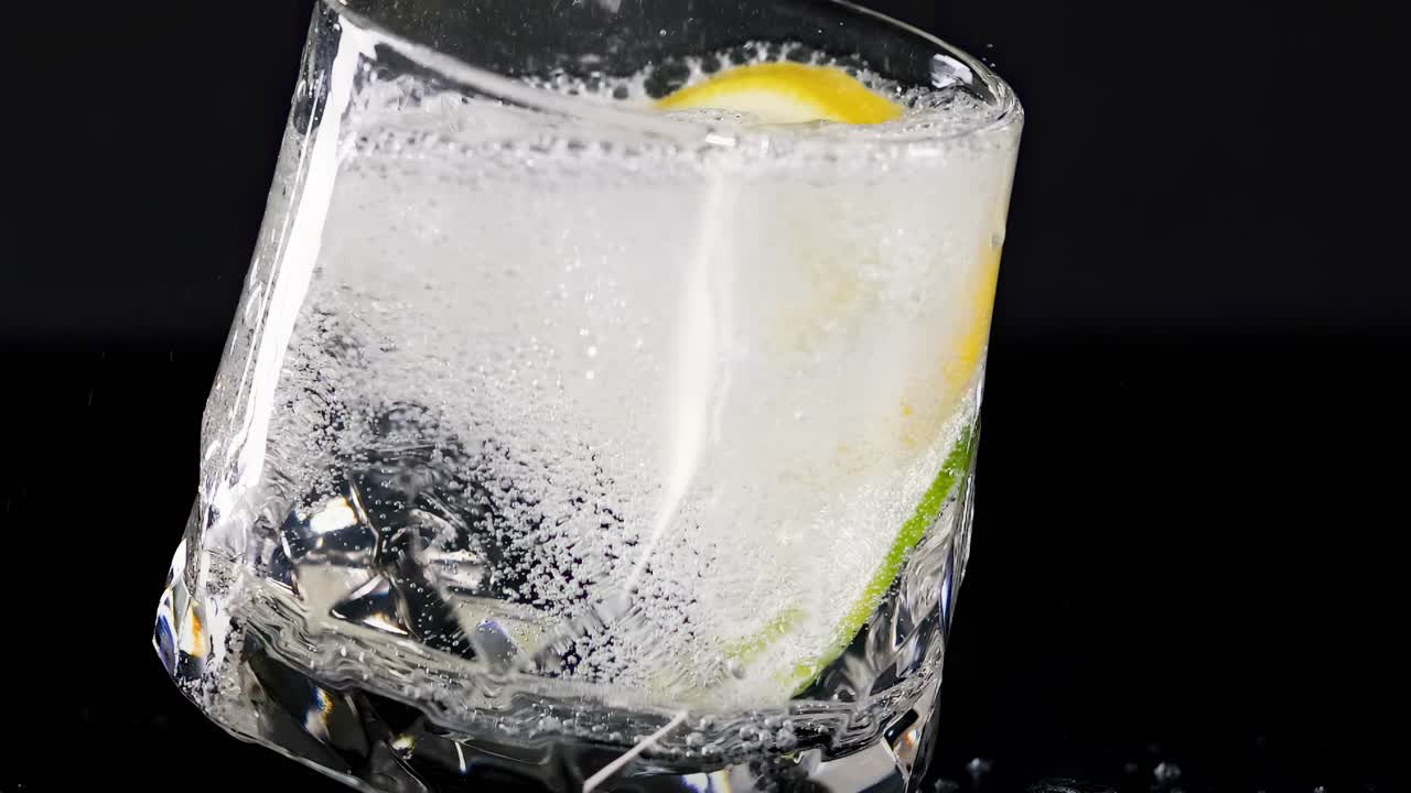 A crystal glass tilts, showcasing a lemon slice and effervescent bubbles against a dark background.