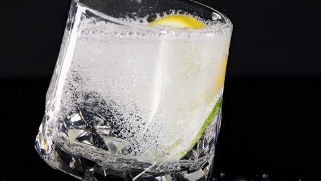 a crystal glass tilts, showcasing a lemon slice and effervescent bubbles against a dark background.