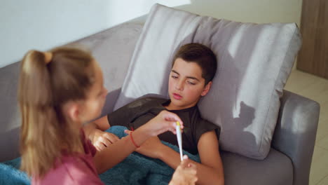 sister with ponytail and maroon shirt gently removes thermometer from mouth of sick brother lying on couch under blue blanket showing care concern and responsibility