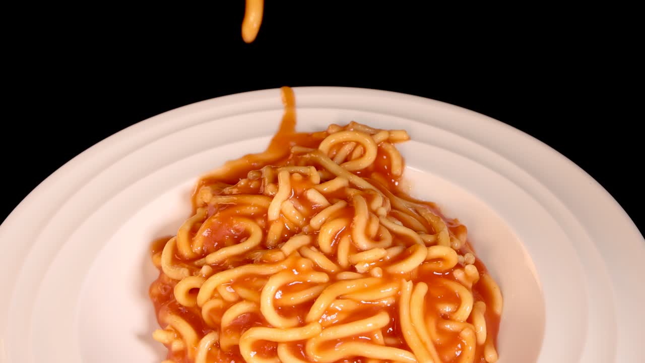 Cooked spaghetti in tomato sauce falls onto a clean white plate against a black background, captured in close-up with bright, even lighting and minimal camera movement