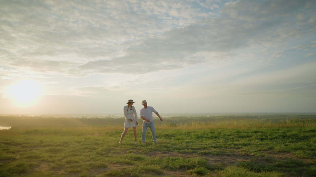 즐거운 부부는 해가 지는 동안 풀이 많은 언덕에서 함께 춤을 추고, 배경에는 조용한 풍경이 있습니다. 색 셔츠와 모자를 입은 남자와 색 드레스를 입은 여자는 손을 놀랍게 움직입니다.
