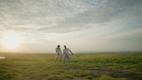 una pareja alegre baila juntos en una colina cubierta de hierba al atardecer, con un paisaje sereno en el fondo. el hombre con una camisa blanca y un sombrero y la mujer con un vestido blanco fluido mueven sus manos juguetonamente