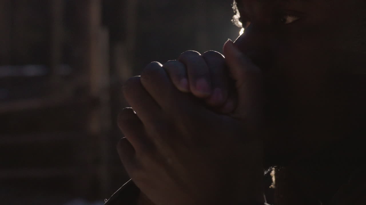 차가운 황혼 속에서 손을 녹이는 흑인 남성, 입가에 모은 손바닥으로 입김이 보임, 도시의 나무 울타리와 림라이트, 조용한 고독과 회복력, 분위기 있는 어두운 조명과 실질적인 생존