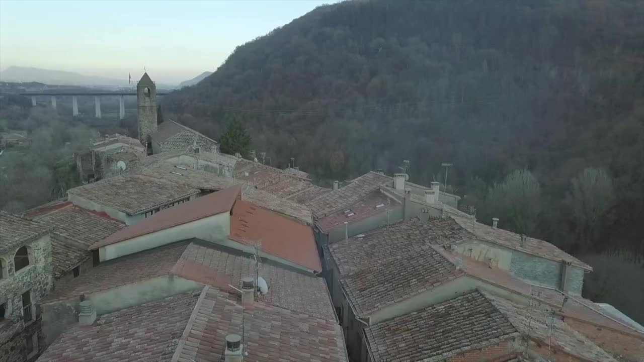 Filmato cinematografico a scorrimento orizzontale di Castellfollit de la Roca, con l'antica torre campanaria della chiesa e le case in pietra arroccate su una ripida scogliera basaltica che domina una profonda valle fluviale catalana.