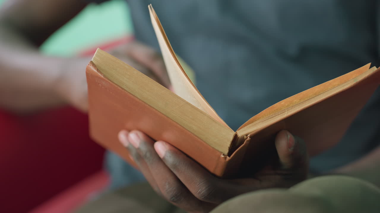 Close up of dark skin person holding worn brown book while reading in relaxed indoor setting, with focus on hands turning pages and warm ambient light creating calm and thoughtful mood