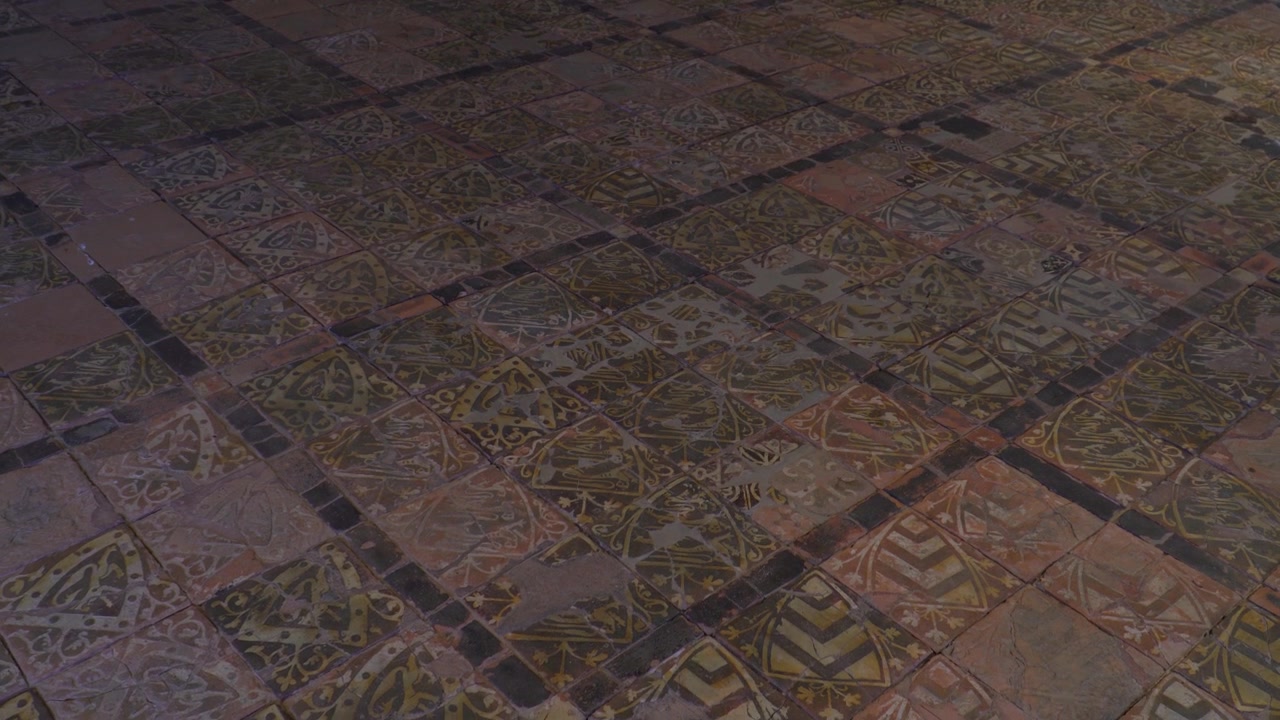 Lenta panoramica cinematografica attraverso piastrelle da pavimento medievali in encausto all'interno dell'Abbazia di Cleeve, che mostra intricati motivi geometrici e artigianato conservato, circondata da pareti in pietra texturizzata.