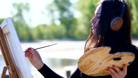 summer, near river, on beach. at sunrise, a beautiful woman artist in big brown headphones holds a brush, palette in her hands, paints on canvas god shiva. listening music, relax