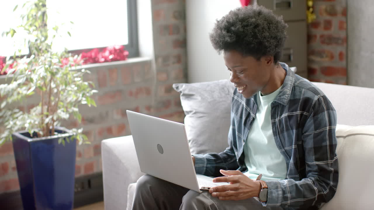 Happy african american man sitting on sofa using laptop at home, slow motion